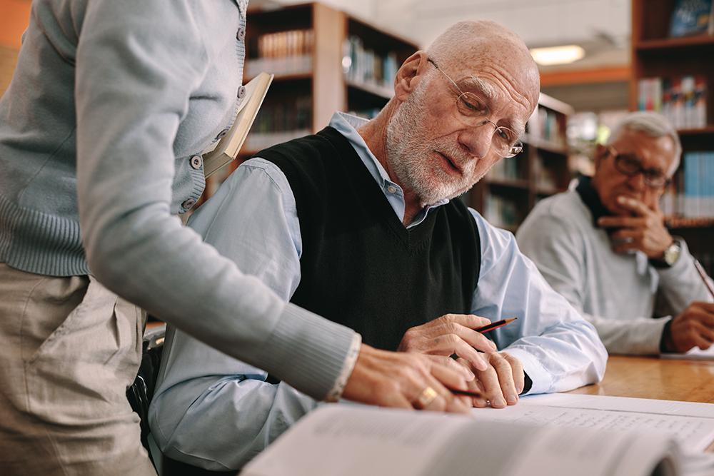 Older man is sitting in a library looking at a book with someone helping them write something down.