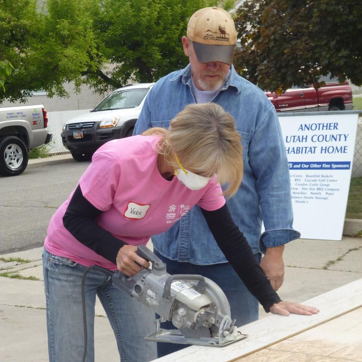 Woman uses a table saw to cut wood while a man wearing a baseball cap watches.